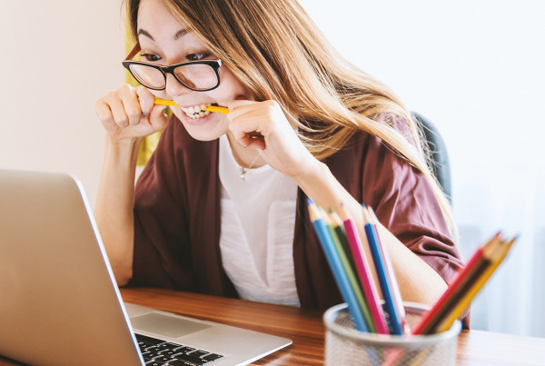En kvinna sitter vid en laptop och biter på en penna för att illustrera att hon är pepp eller frustrerad, fast med ett leende.