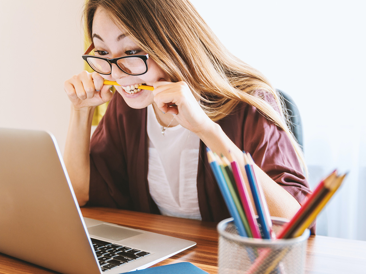 En kvinna sitter vid en laptop och biter på en penna för att illustrera att hon är pepp eller frustrerad, fast med ett leende.